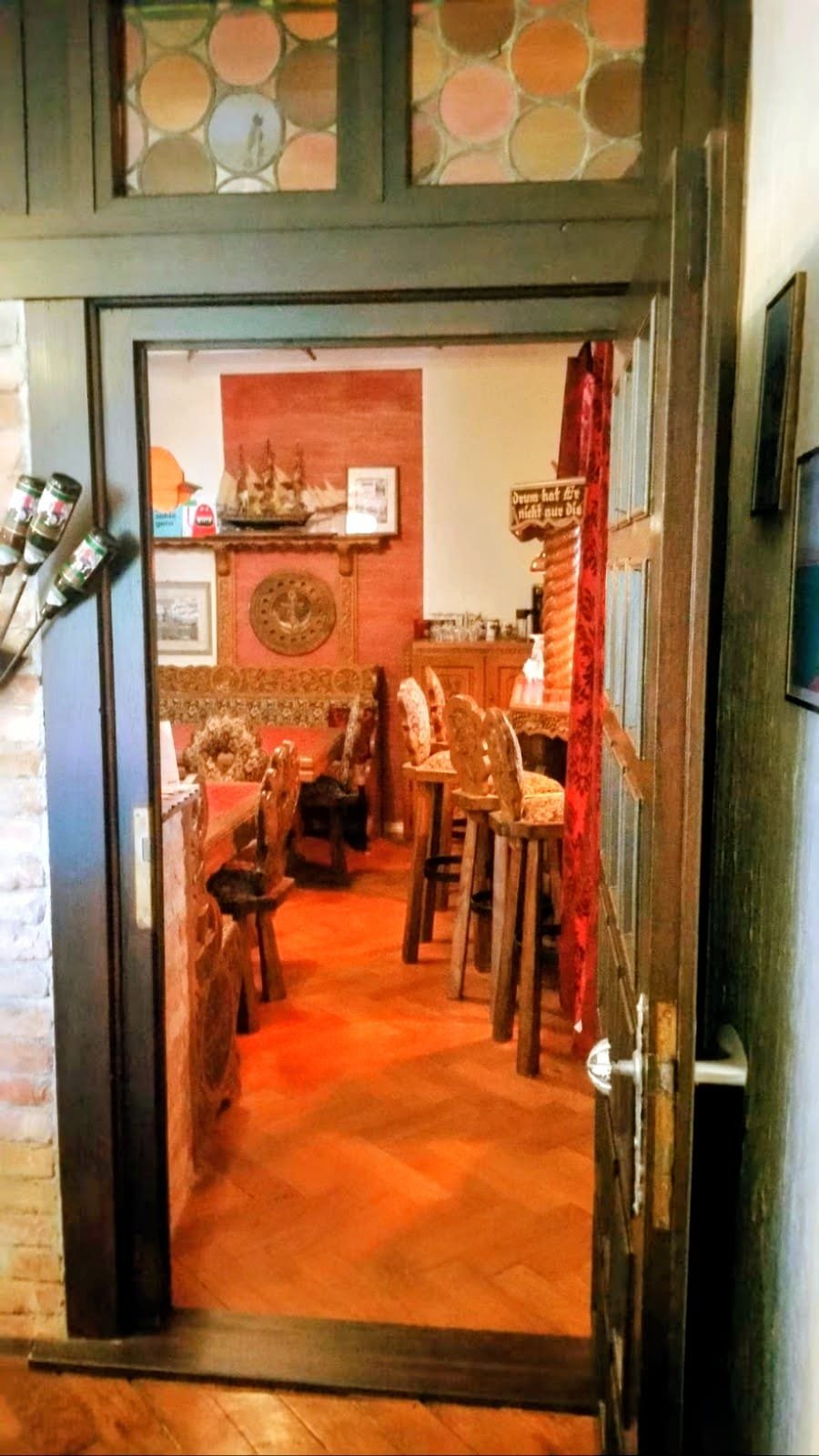 Blick durch die offene Tür in die Hafenkneipe: geschnitzte Holzmöbel, Barhocker, ein Schiffsmodell und ein Anker an der Wand