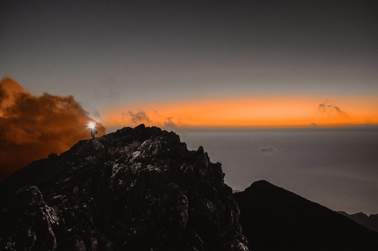 Eine Läuferin mit Stirnlampe auf einem dunklen Felsgrat der Tramuntana, dahinter ein orange glühender Horizont über dem Meer
